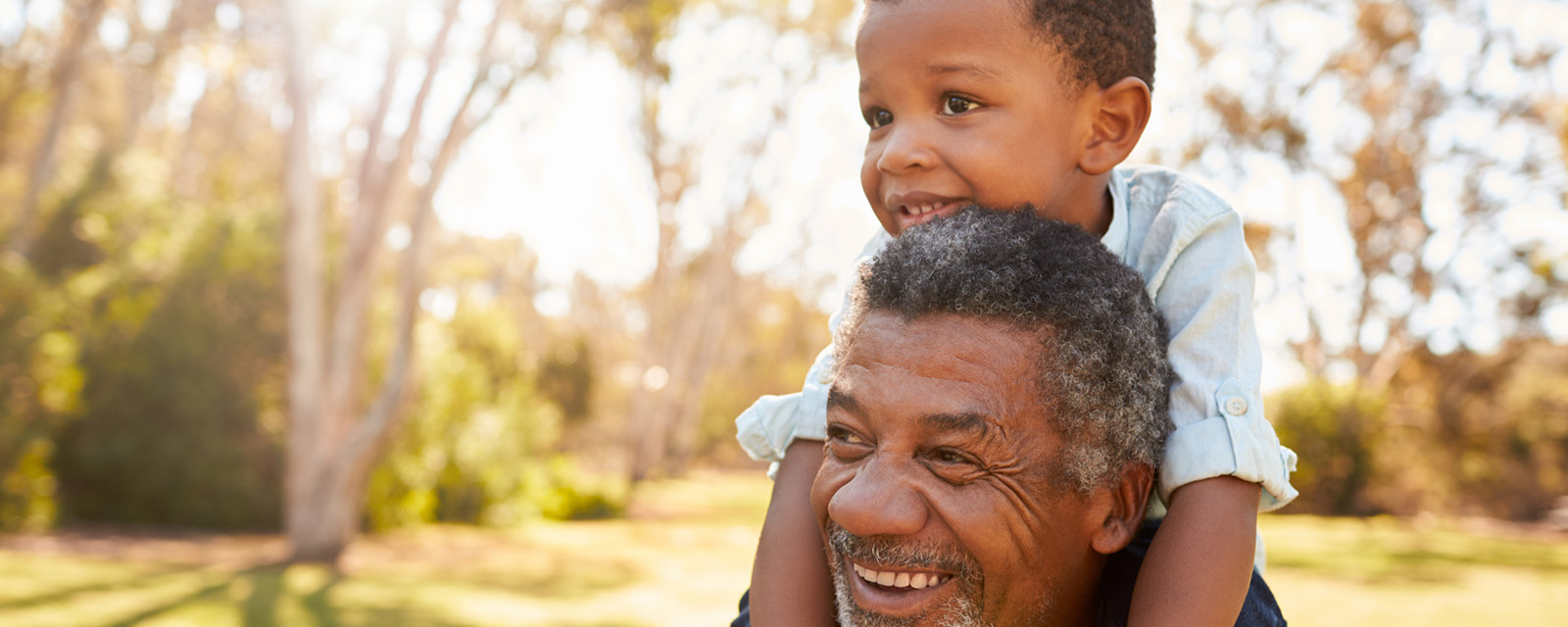 A grandfather carrying his grandson on his shoulders through a park.